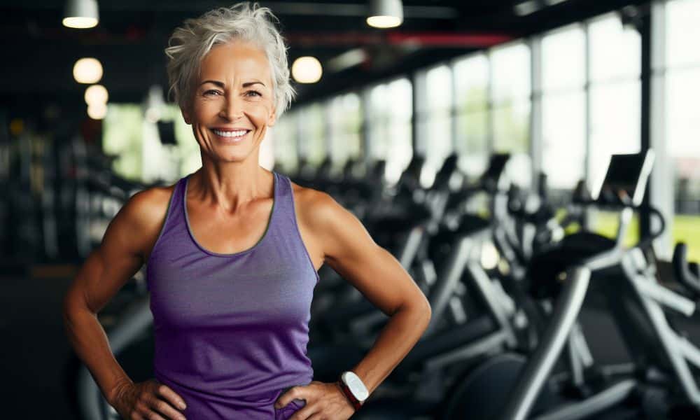 A fit older woman poses in a gym with her hands on her hips.