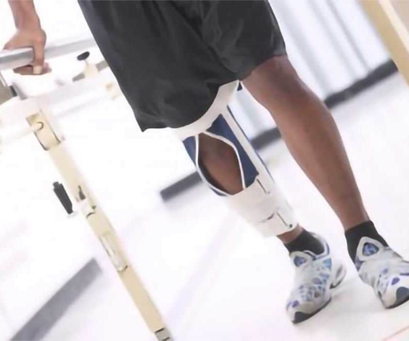 A man with a brace on his leg stands with the support of a bar, during a rehab session.