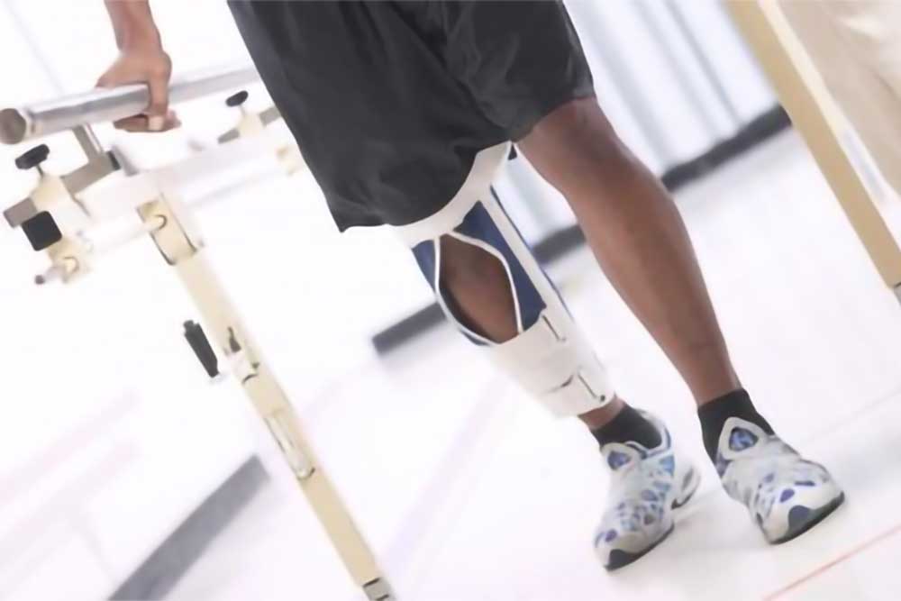 A man with a brace on his leg stands with the support of a bar, during a rehab session.