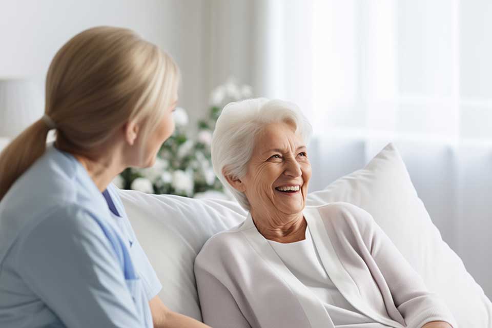 A nurse sits beside an elderly woman propped up on a bed