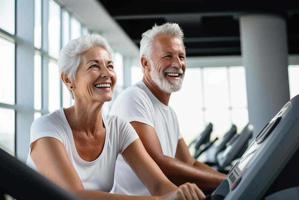 An older couple exercises together on treadmills in a bright gym, smiling and enjoying their workout.