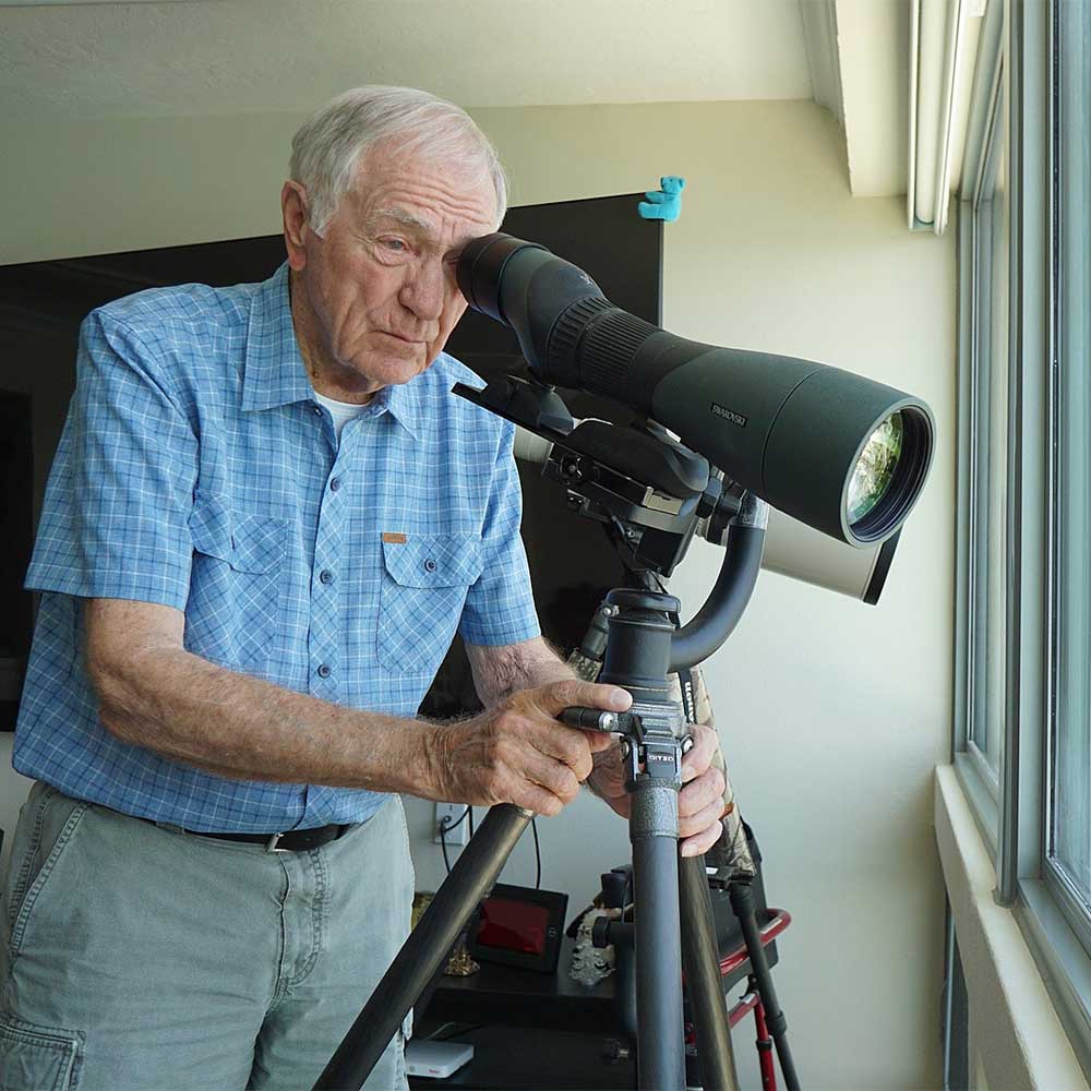 An older man, a wildlife photographer at Plymouth Harbor, gazes through a camera lens, capturing the beauty of nature.
