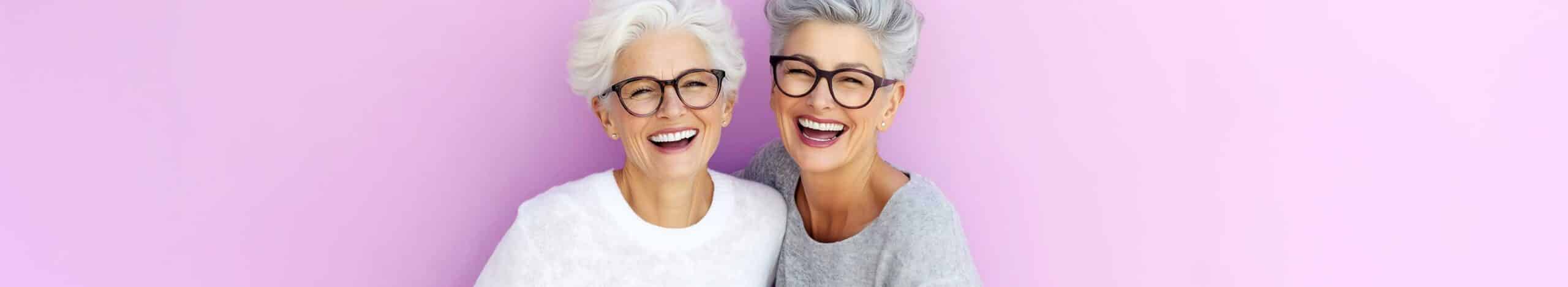 Two senior women with glasses laughing on pink background