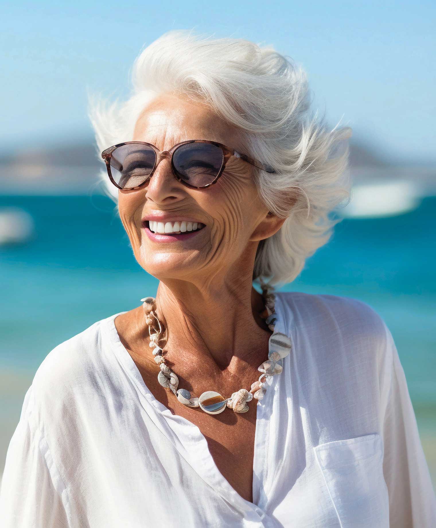 An older woman with white hair and sunglasses enjoys a sunny day at the beach, with waves gently crashing in the background.
